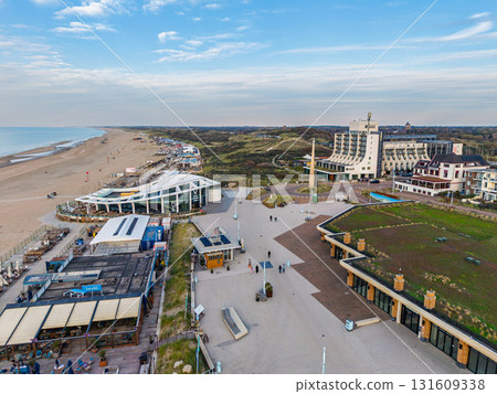 Aerial view of beachfront promenade with modern buildings, green roofed hotel, curved white structure, and sandy shore blending coastal leisure with contemporary architecture. 131609338