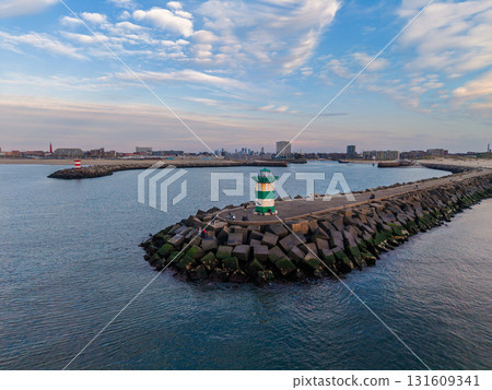 Aerial view of coastal breakwater with green striped lighthouse, angular concrete blocks, and distant cityscape blending maritime infrastructure with urban backdrop under scenic skies. Aerial view of coastal breakwater with green striped lighthouse, angular concrete blocks, and distant cityscape blending maritime infrastructure with urban backdrop under scenic skies. 131609341