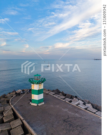 Aerial view of concrete pier with green-striped lighthouse and wave breakers, sailboat in distance capturing serene maritime atmosphere under soft, streaked clouds. 131609342