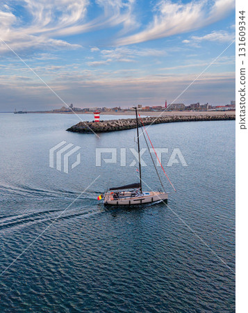 Aerial view of sailboat approaching curved breakwater with red striped lighthouse, calm sea, and urban skyline capturing serene maritime navigation and coastal city backdrop. 131609344