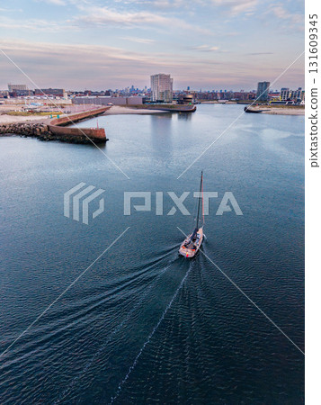 Aerial view of coastal harbor with sailboat leaving wake, flanked by breakwaters and dense city skyline blending maritime movement with urban infrastructure under muted skies. 131609345