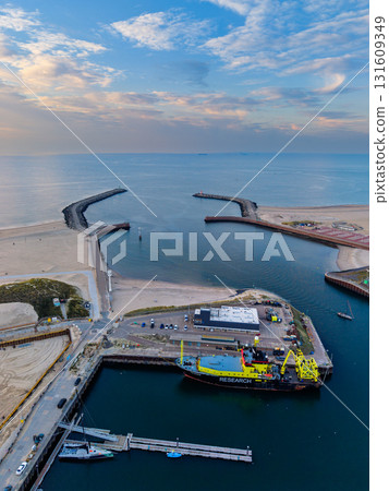 Aerial view of harbor with yellow blue research vessel docked near RESEARCH facility, flanked by breakwaters, sandy beaches, and coastal infrastructure under sunrise or sunset skies. Aerial view of harbor with yellow blue research vessel docked near RESEARCH facility, flanked by breakwaters, sandy beaches, and coastal infrastructure under sunrise or sunset skies. 131609349