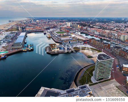 Aerial view of harbor city with curved docks, moored boats, and modern waterfront building framed by dense urban layout and sandy coastline under serene, partly cloudy skies. Aerial view of harbor city with curved docks, moored boats, and modern waterfront building framed by dense urban layout and sandy coastline under serene, partly cloudy skies. 131609350