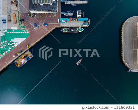 Aerial view of harbor with small boat leaving wake, moored vessels, industrial buildings, and curved breakwater highlighting maritime activity and waterfront infrastructure. 131609351