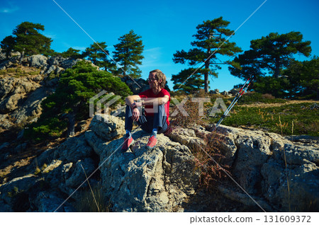 Hiker Resting On Rocky Ridge With Trekking Poles In Sunny Mountain Landscape Hiker Resting On Rocky Ridge With Trekking Poles In Sunny Mountain Landscape 131609372