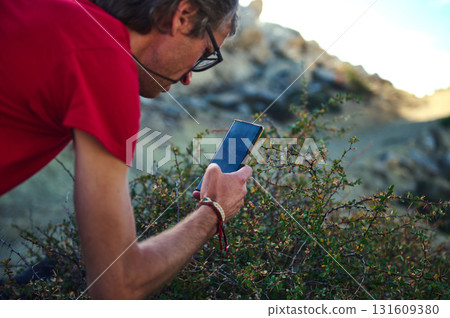 Man In Red Shirt Using Smartphone Outdoors Near Bushes And Rocks 131609380