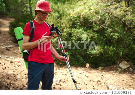 Hiking Enthusiast Checking Phone On Trail With Trekking Poles And Backpack In Sunny Forest 131609381