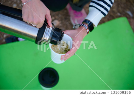 Outdoor Picnic Scene Of A Person Pouring Hot Drink From A Stainless Thermos Into A White Cup On A Bright Green Mat Outdoor Picnic Scene Of A Person Pouring Hot Drink From A Stainless Thermos Into A White Cup On A Bright Green Mat 131609396