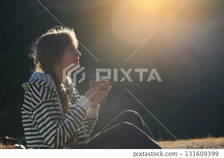 Golden Hour Outdoor Portrait Of A Woman In A Striped Hoodie Sitting On A Hill, Sipping From A Cup, Lost In Thought Amid Warm Sunset Light 131609399