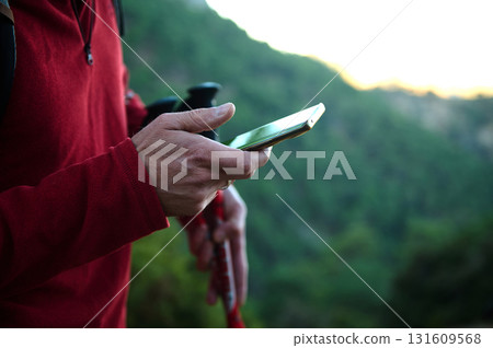 Person In Red Jacket Using Smartphone Outdoors With Hiking Poles In Green Mountain Landscape Person In Red Jacket Using Smartphone Outdoors With Hiking Poles In Green Mountain Landscape 131609568