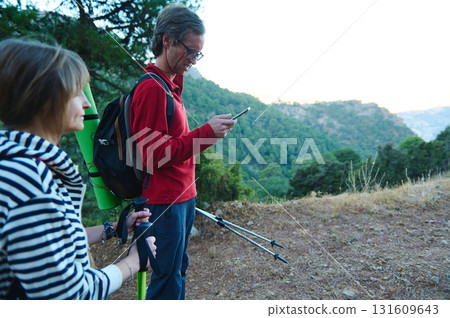 Hiking Friends On a Trail: Man With Red Jacket Looks at Phone While Woman Holds Trekking Poles in Scenic Mountain Outdoors 131609643