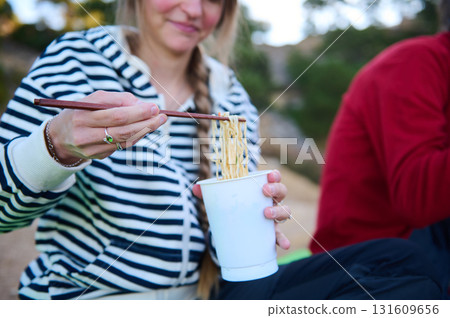 Friends Enjoy Noodles Outdoors: Woman Uses Chopsticks to Lift Noodles From a Paper Cup Friends Enjoy Noodles Outdoors: Woman Uses Chopsticks to Lift Noodles From a Paper Cup 131609656