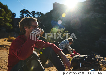 Outdoor Photography Scene With Man Resting And Sunlit Mountain Landscape As Friend Packs Gear, Capturing Natural Light And Calm Moment During Hike 131609660