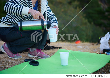 Outdoor Picnic Moment: Woman Pouring From Thermos Into Cup On Green Mat During Outdoor Adventure Outdoor Picnic Moment: Woman Pouring From Thermos Into Cup On Green Mat During Outdoor Adventure 131609712