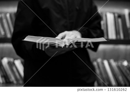 Person is holding open book in library. Person appears to be reading or studying the book. The library is filled with numerous books. Concept of learning and self development in religion Person is holding open book in library. Person appears to be reading or studying the book. The library is filled with numerous books. Concept of learning and self development in religion 131610262