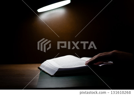 Hand gently resting on pages of open book, illuminated by focused light source, creating serene and contemplative atmosphere. Emphasizing details of book and hand 131610268