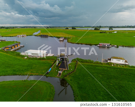 Aerial view of Dutch countryside with central windmill, houseboats, canal, and green fields capturing iconic rural charm and waterway culture under dramatic overcast skies. 131610565