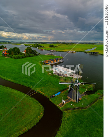 Aerial view of Dutch countryside with central windmill, houseboats, canal, and green fields capturing iconic rural charm and waterway culture under dramatic overcast skies. Aerial view of Dutch countryside with central windmill, houseboats, canal, and green fields capturing iconic rural charm and waterway culture under dramatic overcast skies. 131610566