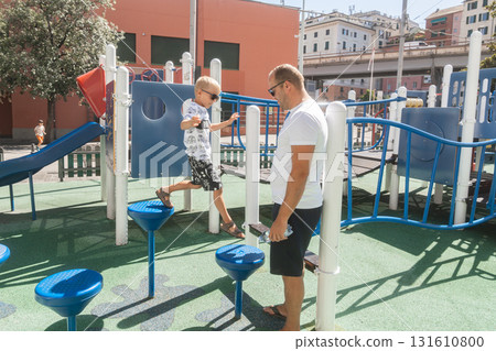 Father watching his son play on playground equipment. Family bonding, leisure, parenthood, and active lifestyle outdoors. Father watching his son play on playground equipment. Family bonding, leisure, parenthood, and active lifestyle outdoors. 131610800