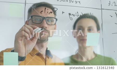 Professional team members brainstorming strategy, writing notes on transparent whiteboard with colorful sticky papers during collaborative meeting in office. Business people at work Professional team members brainstorming strategy, writing notes on transparent whiteboard with colorful sticky papers during collaborative meeting in office. Business people at work 131612340