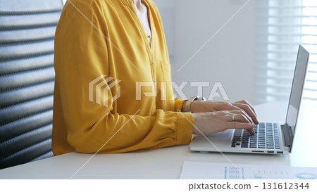 Unknown businesswoman wearing yellow blouse working on laptop at minimalist white desk in contemporary workplace, typing with focus. Business people concept Unknown businesswoman wearing yellow blouse working on laptop at minimalist white desk in contemporary workplace, typing with focus. Business people concept 131612344