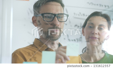 Professional businesspeople collaborating, reviewing data on transparent whiteboard, developing strategic corporate solutions in modern collaborative workspace. Business people concept 131612357