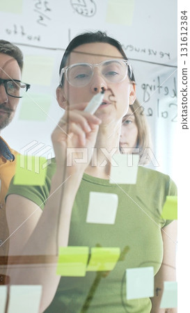 Professional team brainstorming, sharing strategy on glass whiteboard with colorful sticky notes, working collaboratively in bright office space 131612384