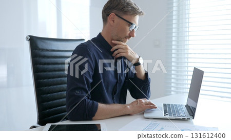 Businessman sitting at a sleek desk, working on a laptop and thinking in a bright, modern office filled with natural light. Business people concept Businessman sitting at a sleek desk, working on a laptop and thinking in a bright, modern office filled with natural light. Business people concept 131612400