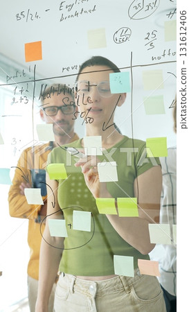 Adult casual dressed businesswoman wearing glasses writing on glass board, marking strategic plan with sticky notes while colleagues brainstorming in background. Designers at work 131612406