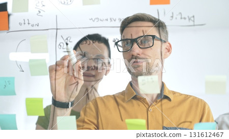 Business colleagues brainstorming, writing strategy notes on glass board, collaborating with colorful sticky notes in creative workplace meeting Business colleagues brainstorming, writing strategy notes on glass board, collaborating with colorful sticky notes in creative workplace meeting 131612467