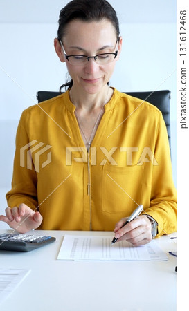 Businesswoman wearing yellow shirt is sitting at white desk and using calculator while taking notes, with laptop and folders in the background. Audit and taxes in business 131612468