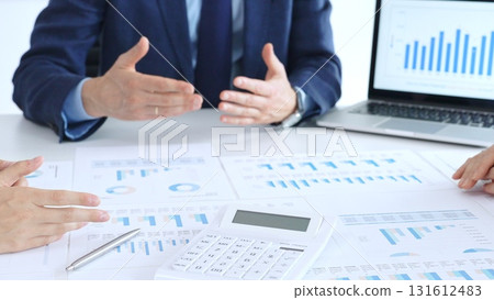 Business professionals collaborating at a conference table, reviewing financial charts and reports, using a laptop and calculator while engaging in discussions about strategy and performance 131612483