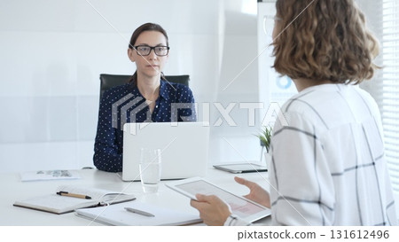 Professional women collaborating at white workspace, reviewing digital content on tablet and laptop during strategic work session 131612496