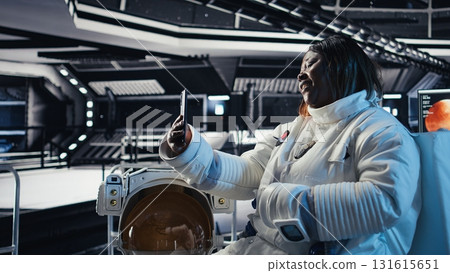 Happy female cosmonaut doing videocall with family using smartphone during galaxy expedition. Africna american woman doing remote call with loved ones using mobile phone in spaceship Happy female cosmonaut doing videocall with family using smartphone during galaxy expedition. Africna american woman doing remote call with loved ones using mobile phone in spaceship 131615651