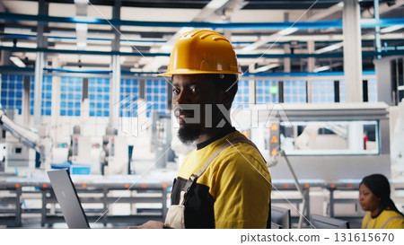 Photovoltaics factory technician optimizing production flow using laptop, wearing hardhat. Sustainable energy solar panels plant specialist monitoring manufacturing process on device. Camera B. 131615670