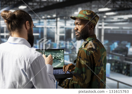Military technician in data center monitoring warfare drone imagery in server farm command center. IT specialist in army server room using tactical interface to support aerial operations. Military technician in data center monitoring warfare drone imagery in server farm command center. IT specialist in army server room using tactical interface to support aerial operations. 131615763