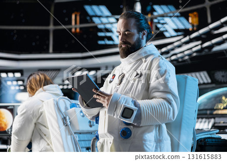 Astronaut wearing spacesuit inside futuristic spacecraft monitoring mission data on tablet screen. Spaceman wearing equipment helmet prepares for space travel operations, reading data on device 131615883