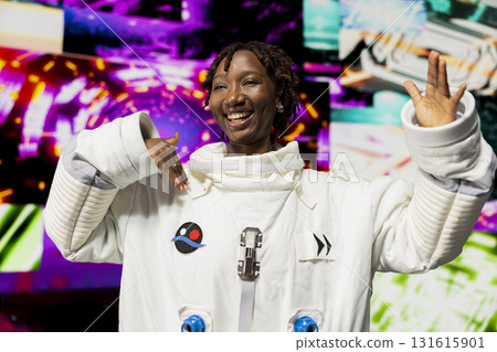 Joyous woman wearing astronaut suit doing dance moves with hands reacting to abstract colorful visuals on digital background. Upbeat cosmonaut having fun, isolated against bright neon vortex Joyous woman wearing astronaut suit doing dance moves with hands reacting to abstract colorful visuals on digital background. Upbeat cosmonaut having fun, isolated against bright neon vortex 131615901