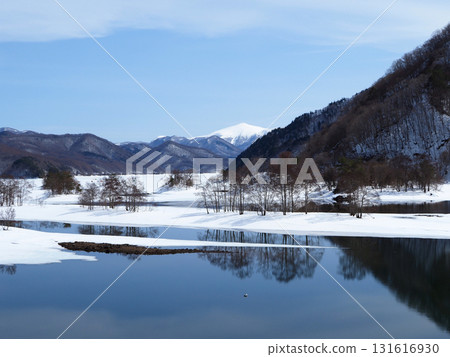 Lake Akimoto in Urabandai in winter. The ice has melted, and the snow-capped mountains are strikingly beautiful against the white snow fields and the trees reflected on the calm lake surface. Lake Akimoto in Urabandai in winter. The ice has melted, and the snow-capped mountains are strikingly beautiful against the white snow fields and the trees reflected on the calm lake surface. 131616930