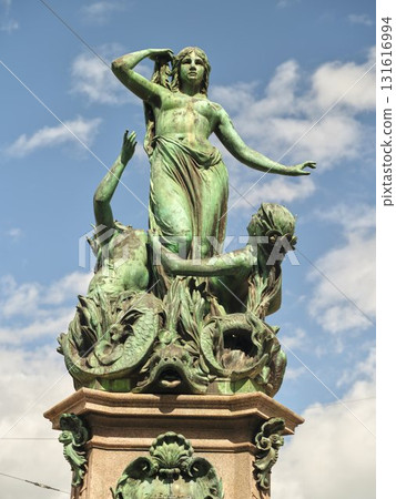 Bronze statue depicting a woman with flowing hair and expressive pose surrounded by mythological figures in a clear blue sky 131616994