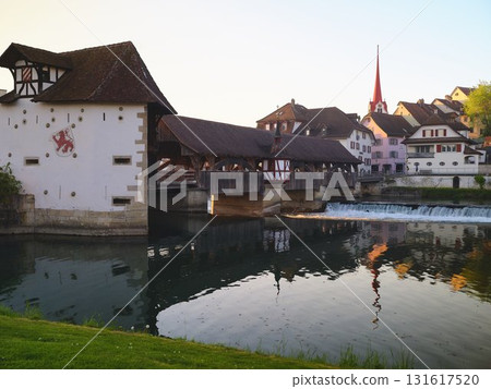Historic wooden bridge and charming buildings reflect in calm water during golden hour in picturesque Swiss town Historic wooden bridge and charming buildings reflect in calm water during golden hour in picturesque Swiss town 131617520
