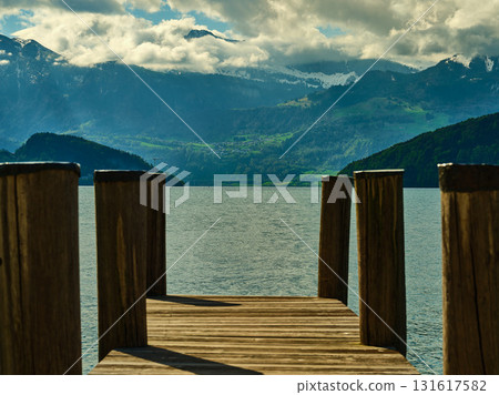 Dock extending over tranquil lake with mountains in the background during a cloudy day 131617582