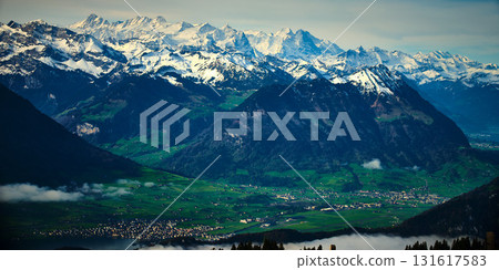 Majestic snow-capped mountains rise above lush green valleys in a picturesque landscape during a clear daytime sky 131617583