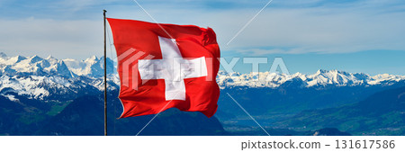 Majestic Swiss flag waving against the backdrop of snow-capped mountains and clear blue skies Majestic Swiss flag waving against the backdrop of snow-capped mountains and clear blue skies 131617586