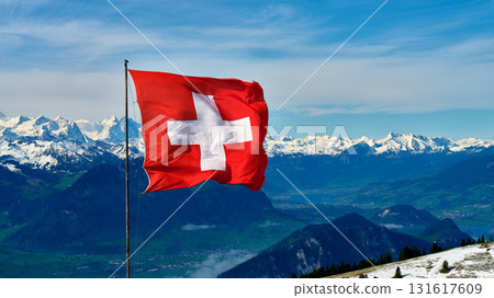 Swiss flag waving proudly in the mountains with snowy peaks and blue skies in the background 131617609