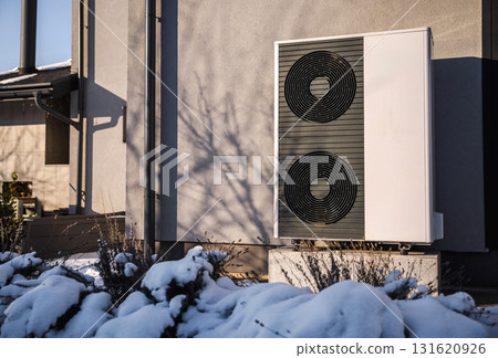 Modern air source heat pump installed outside a residential building in winter. Snow-covered plants and sunlight reflect the concept of sustainable home heating and energy efficiency. Modern air source heat pump installed outside a residential building in winter. Snow-covered plants and sunlight reflect the concept of sustainable home heating and energy efficiency. 131620926