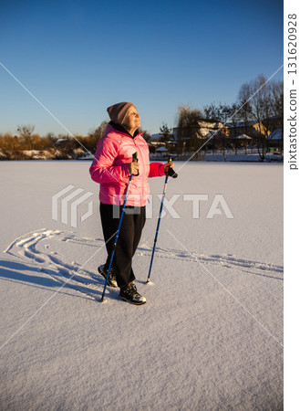 Elderly woman walking with trekking poles on a snowy field during sunny winter day. She wears a pink jacket and smiles, enjoying outdoor exercise and fresh air in the cold season. 131620928