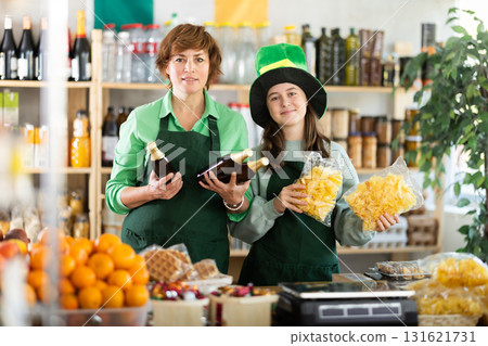 Two female sellers posing in green costume and hat, offering bottle of irish ale in store decorated for St. Patrick Day Two female sellers posing in green costume and hat, offering bottle of irish ale in store decorated for St. Patrick Day 131621731