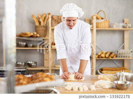 Young guy baker in white uniform cooking bread in kitchen, kneading dough on table 131622178