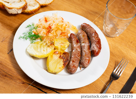 On table there is plate with grilled sausages, sauerkraut and slices of potatoes 131622239